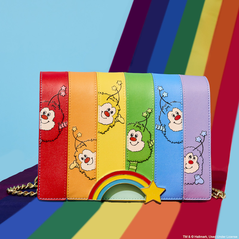 Loungefly Rainbow Brite Rainbow Sprites Crossbody Bag sitting on a rainbow against a blue background. The bag features vertical columns that are each a different color of the rainbow, with that color Sprite in the column. 
